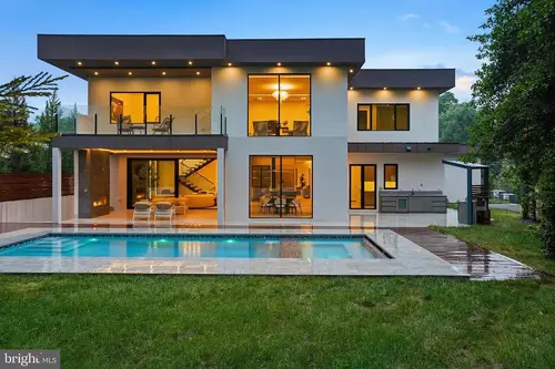 The back of the house at dusk — a flat-roofed white modern facade under deep dark eaves, a glass-railed terrace over the lit living floor, and a pool across the stone patio