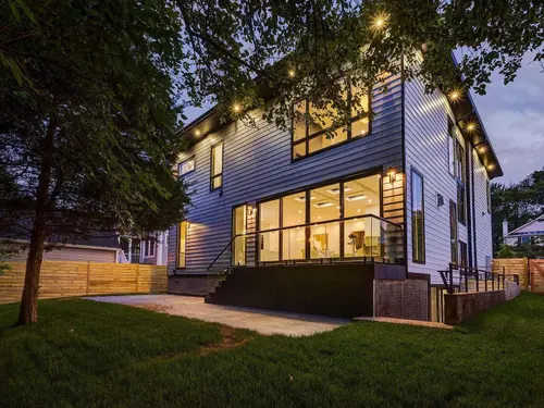 The house from the garden at dusk — pale lap siding and black window frames, a raised terrace behind a glass balustrade, and steps down to the paved patio