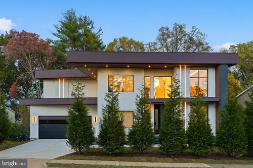The front of the house at dusk — white stucco under wide dark flat-roof overhangs, vertical light strips, and a row of evergreens along the entrance