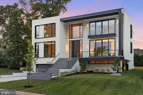 The front of the house at dusk — a three-story white modern facade with dark trim, glass-railed balconies and a broad stone stair up to the door