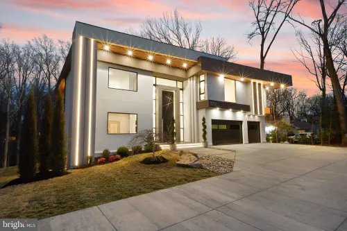 The house at dusk — a flat-roofed modern facade in white stucco with lit vertical reveals and a wood soffit