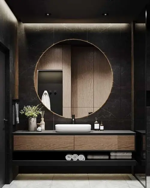 A round mirror on a dark stone wall above an oak vanity, with a lit shelf of towels below it