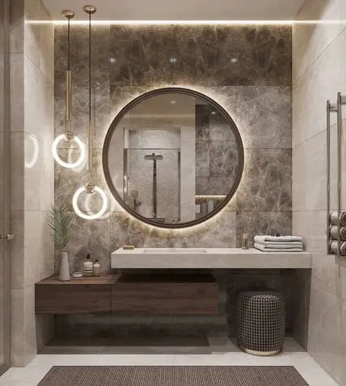 A round backlit mirror on a marble wall over a stone basin shelf, with a walnut drawer unit beneath and ring pendants beside it